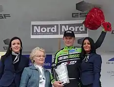 De face, quatre personnes sourient. Un homme habillé en noir et vert lève un bouquet, tout en tenant un trophée.