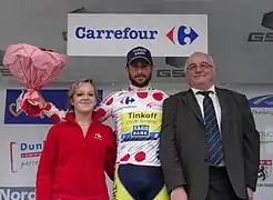 De face, trois personnes sont alignées. Un homme avec un maillot blanc à pois rouges lève un bouquet de fleurs rouge.