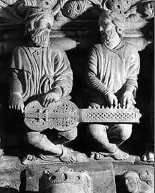 Photo de sculpture représentant les deux instrumentistes d'un organistrum, l'un à la manivelle, l'autre aux tirettes