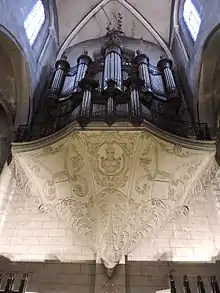 Photo d'un orgue en position haute, sous la voute d'une église. Il est supporté par un  piédestal ouvragé.