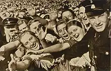 Photographie en noir et blanc d'une foule joyeuse, contenue par des hommes en uniforme sombre.