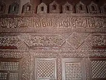 Photographie de la partie supérieure de la maqsura, montrant divers ornements sculptés, bandeau épigraphique et merlons ajourés.