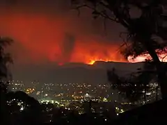 Photographie le soir d'un village dans une vallée. La ligne d'horizon dans les collines au-delà est éclairée en rouge par des feux de forêt.