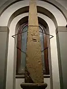 Photographie d'une stèle/obélisque en pierre assez imposante, exposé devant une fenêtre, avec un bas-relief en très mauvais état au milieu.