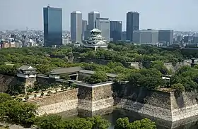 Photographie où l'on voit des remparts au premier plan, la tour principale au second, et des hautes tours modernes à l'arrière-plan.