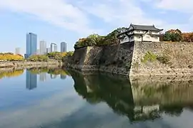Photographie montrant des remparts de plusieurs mètres de haut entourés d'eau. Des tours modernes sont visibles à l'arrière-plan.