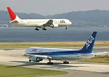 Un appareil de la Japan Airlines, de couleur blanche avec la dérive rouge, au-dessus de la piste avec le train d'atterrissage sorti ; un avion fr All Nippon Airlines dans une livrée bleue et blanche est au roulage.
