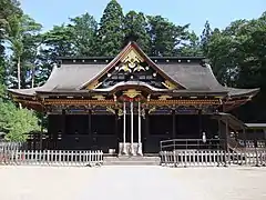 Photo couleur de la façade ombragée de la salle du culte d'un sanctuaire shintō, structure en bois foncé dont le frontispice est doré par endroits. L'arrière-plan est constitué d'une rangée d'arbres au feuillage vert sous un ciel bleu sans nuages.