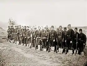 Photographie d'une rangée de soldats ottomans en uniforme, fusil au côté et posant pour le photographe.