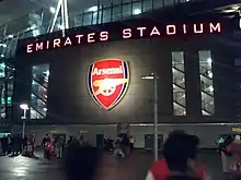 Photo de l'entrée du stade de nuit avec « Emirates Stadium » en majuscules éclairées sur la façade.
