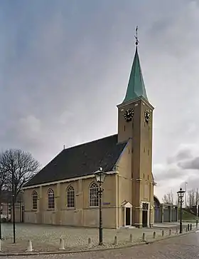 Photographie en couleurs et de trois quarts d'un édifice en briques et de son parvis longé par une rue.