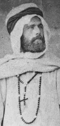 Photographie ancienne d'un jeune homme barbu avec un vêtement blanc et une croix autour du cou.