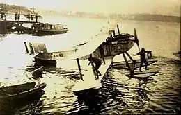 Biplan monomoteur à flotteurs, stationné sur l'eau, avec un équipier sur le flotteur et un autre sur l'aile inférieure.