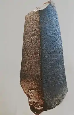Photographie d'une stèle pyramidale à base rectangulaire, en pierre foncée.  Les deux faces visibles sont gravées de nombreuses inscriptions.