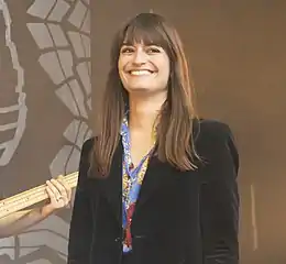 Femme blanche jeune avec les cheveux longs châtains, photographiée de face, souriante ; le manche d'une basse et la main du bassiste est visible dans l'arrière-plan à gauche.