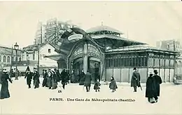 Photo sépia d'une esplanade avec des gens en manteaux sombres, robes longues et chapeaux devant un édifice à la toiture et à l'entrée tarabiscotées