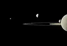Image en noir et blanc où les anneaux externes sont au centre de l'image. Une moitié de Rhéa est montrée ainsi que des croissants des autres lunes.