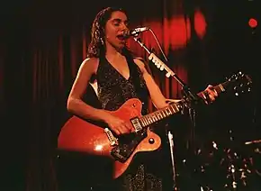 Image en couleur d'une femme brune habillée d'une robe de soir chantant dans un micro et jouant de la guitare