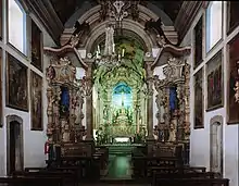 Photographie en couleurs. Intérieur de la nef : les murs sont blancs et affublés de nombreux tableaux ; les angles sont forces de reliefs en couleurs, très baroques, tandis que l'autel au fond semble illuminé d'une lueur verte.
