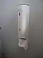 Distributeur vertical de papier toilette, avec déroulement du rouleau dans le sens horaire.