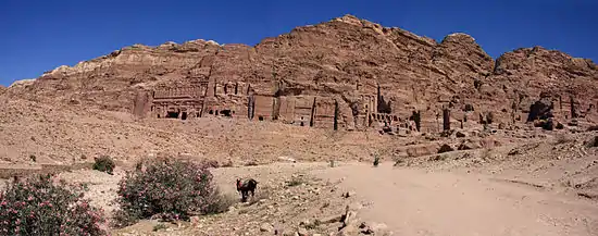 Vue panoramique sur des monuments sculptés dans la roche. À l'avant plan, un chemin qui mène vers les monuments. Sur chemin arrivent deux personnes sur un cheval. Un âne stationne à gauche du chemin.