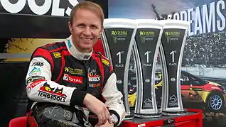 Photographie d'un homme souriant, vu de face avec des trophées derrière lui.