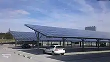 Deux rangées de panneaux photovoltaïques situés en hauteur, avec deux voitures garées sur le parking dessous.