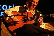 Photographie en plan rapproché d'un homme dans la maturité, assis, jouant de la guitare les yeux mi-clos, la guitare posée sur son genou droit, jambe repliée posée sur genou gauche, dans la posture habituelle pour la guitare flamenca.