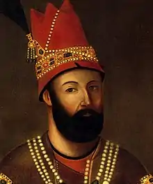 Tableau représentant un homme à la barbe et moustache noire en costume d'apparat, il porte un collier de perles à son cou et un haut chapeau rouge orné de perles et pierres précieuses