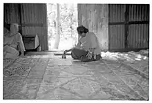 Photo en noir et blanc d'une femme assise en arrière plan dans une grande pièce. Sur le sol s'étend un très grand tissu rempli de motifs géométriques. Une autre femme, assise sur le côté, la regarde.