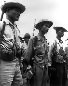 Trois soldats en uniforme avec un chapeau, se tenant debout. Ils sont armés de fusils et grenades. D’autres soldats en arrière-plan.