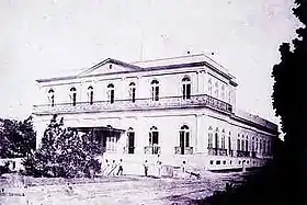 Photographie en noir et blanc d'une grande bâtisse de style néo-classique, surmontée d'un étage coiffé d'un fronton triangulaire et entouré d'un balcon, couvrant en partie le rez-de-chaussée.