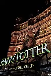 Vue sur la façade d'un bâtiment rouge éclairé de nuit. Au-dessus de l'entrée du bâtiment est placé un décor comportant un nid avec deux ailes et au-dessous duquel se trouve écrit en grosses lettres jaunes « Harry Potter and the Cursed Child »
