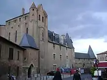 la photo couleur montre un bâtiment en brique rouges, encadrement de fenêtre en pierre et toiture en ardoise. À gauche, on distingue les vitraux d'une chapelle ; à droite, les larges fenêtres à meneau donnent sur le palais