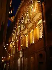 Vue en contre-plongée de la façade d'un palace éclairée de nuit dans une petite rue avec une guirlande d'ampoules zigzaguant entre les murs.