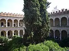 Image photographique avec au centre de la verdure et un arbre qui coupe un édifice en deux en arrière plan dont la façade est composée d'arches sur deux niveaux.