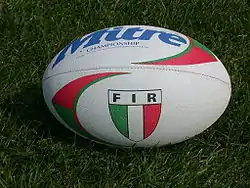 Photographie d'un ballon de rugby de couleur blanc, de marque Mitre, avec un drapeau de l'Italie inscrit dessus.