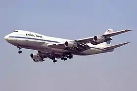 Photo du côté gauche, légèrement de face, d'un avion en vol aux couleurs blanches et bleues de Pan Am avec le train d'atterrissage sorti.