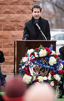 Photo d'un homme se tenant derrière un pupitre avec micro et faisant un discours. Il se tient juste devant le monument commémoratif en pierre et une couronne de fleurs est fixée devant le pupitre.