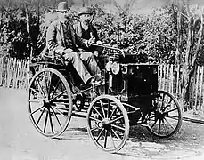 Photo d'un véhicule motorisé de profil conduit par deux hommes, le véhicule ressemble fortement à un véhicule hyppomobile.