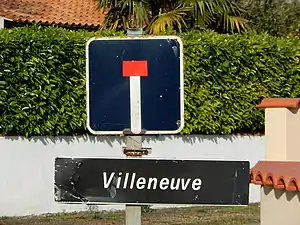 Un panneau C13a surmontant un panneau E31 sur lequel il est écrit le nom du lieu-dit « Villeneuve ».