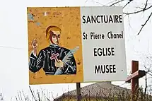 Panneau avec une image d'un homme habillé en noir, tenant une croix et une palme dans la main gauche, le doigt levé dans la main droite ; à droite du panneau, est écrit sur fond blanc "Sanctualre St Pierre Chanel - église - musée"