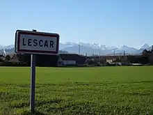 Photographie en couleurs d'un panneau avec l'inscription " Lescar", en fond des montagnes enneigées.