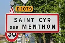 Panneau blanc avec un liseré rouge où il est inscrit "Saint-Cyr-sur-Menthon" en son centre en lettres noires. Ce panneau est surmonté d'un panonceau jaune où il est inscrit "D 1079".