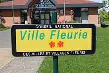 Panneau jaune à liseré noir où il est inscrit "Ville fleurie" avec deux fleurs rouges représentant les fleurs de récompense au concours des villes et villages fleuris