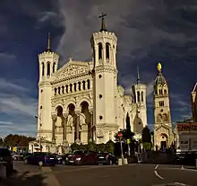 Photographie couleur de la basilique vue de l'angle sud-ouest du parvis : façade occidentale, trois des clochers et statue de la Vierge.