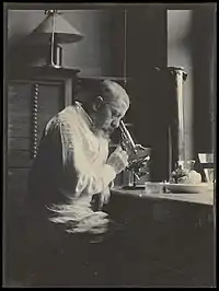 Photographie en noir et blanc d'un homme en blouse blanche penché sur son microscope dans la pénombre, un cerveau humain posé à côté de lui