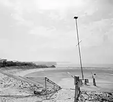 Photographie noir et blanc d'un mat métallique sur une surface parabolique en béton. Un homme se tient à côté du mât.