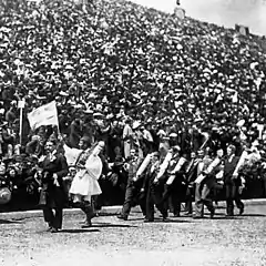 Un homme porte un drapeau grec qu'il monte au public, suivi par d'autres personnes en costume.