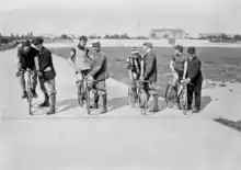Quatre cyclistes sur leur vélo au départ d'une course sur un vélodrome, chacun tenu par un homme debout.
